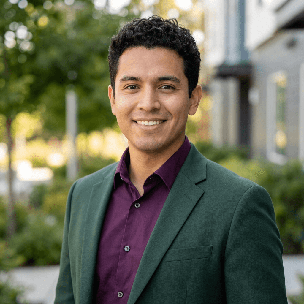 Smiling man with dark curly hair wearing a green blazer and purple shirt outdoors.