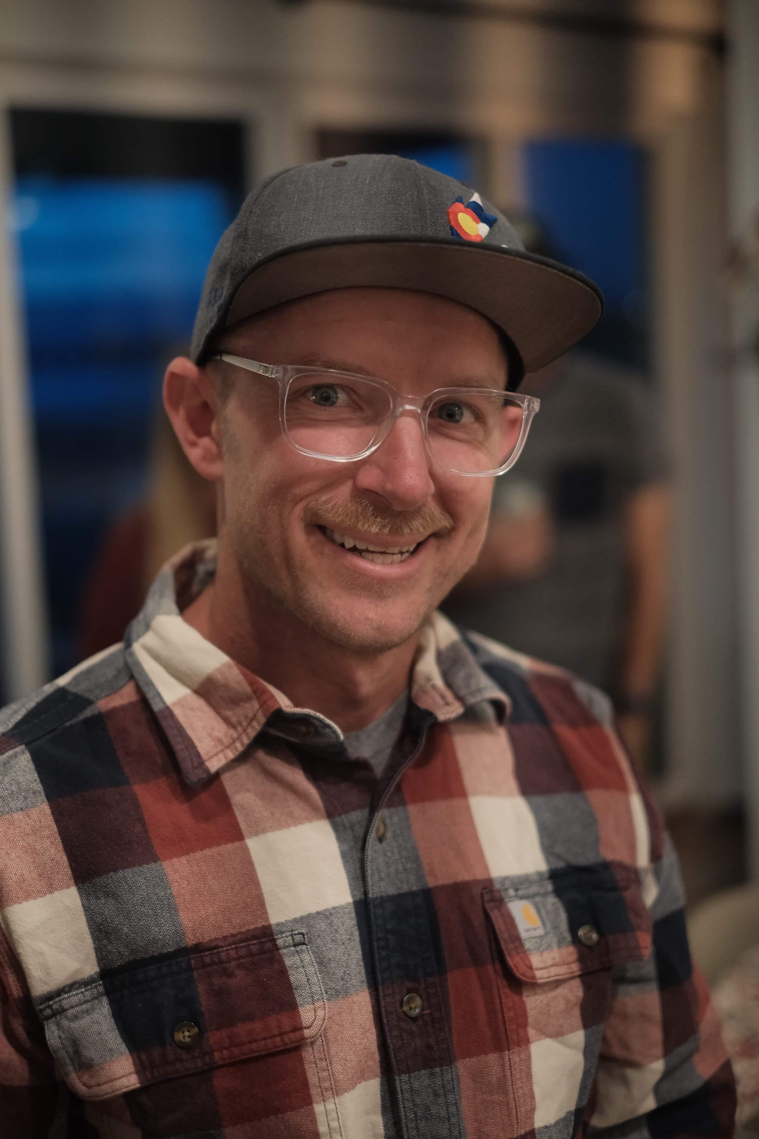 Smiling man with a mustache, clear glasses, grey cap, and red and blue plaid shirt.