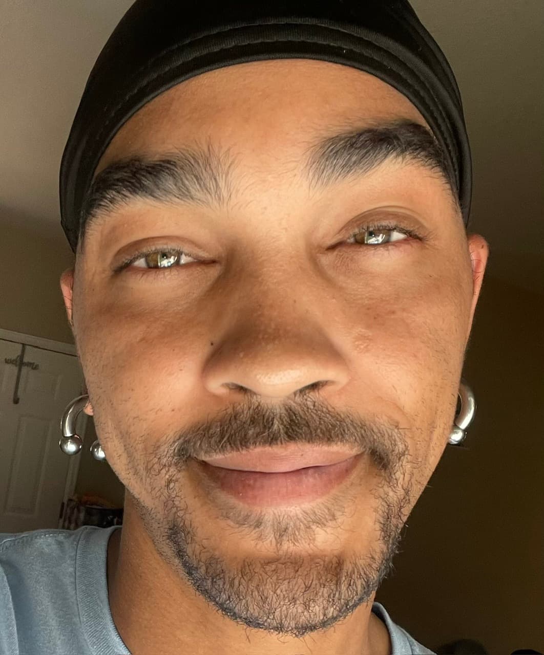 Close-up of a man with hazel eyes, goatee, black durag, and silver hoop earrings.